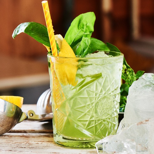 Cóctel alcohólico Basil Smash en una probeta de vidrio con un trozo de hielo sobre una mesa de madera.