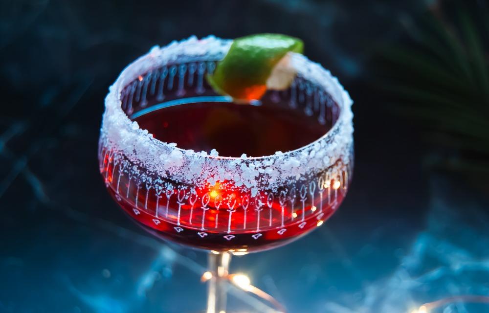 Cocktail rosso alcolico, guarnito con lime, bordo salato, sfondo marmorizzato, bicchiere invecchiato, luci magiche.