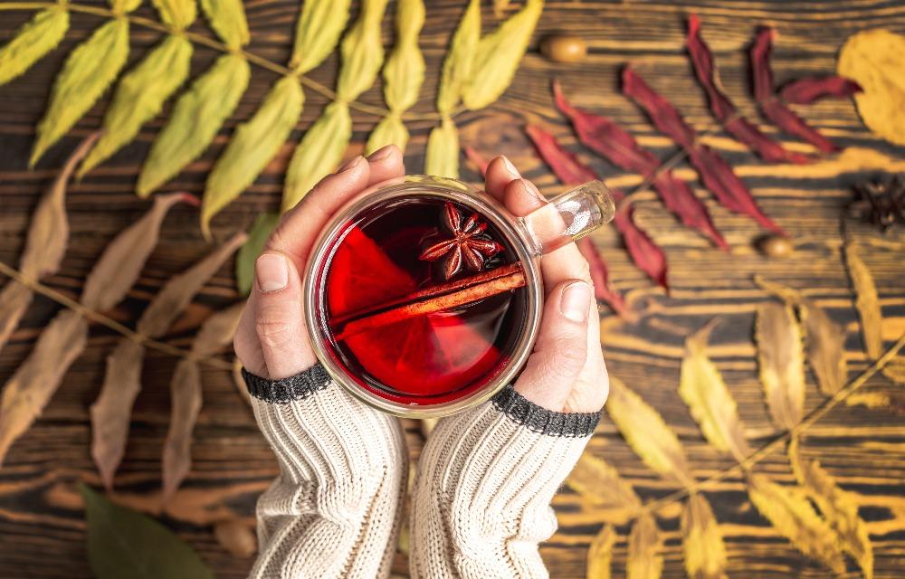 Woman's hands in a sweater holding a glass mug containing a hot drink with cinnamon, orange, and star anise on a background.