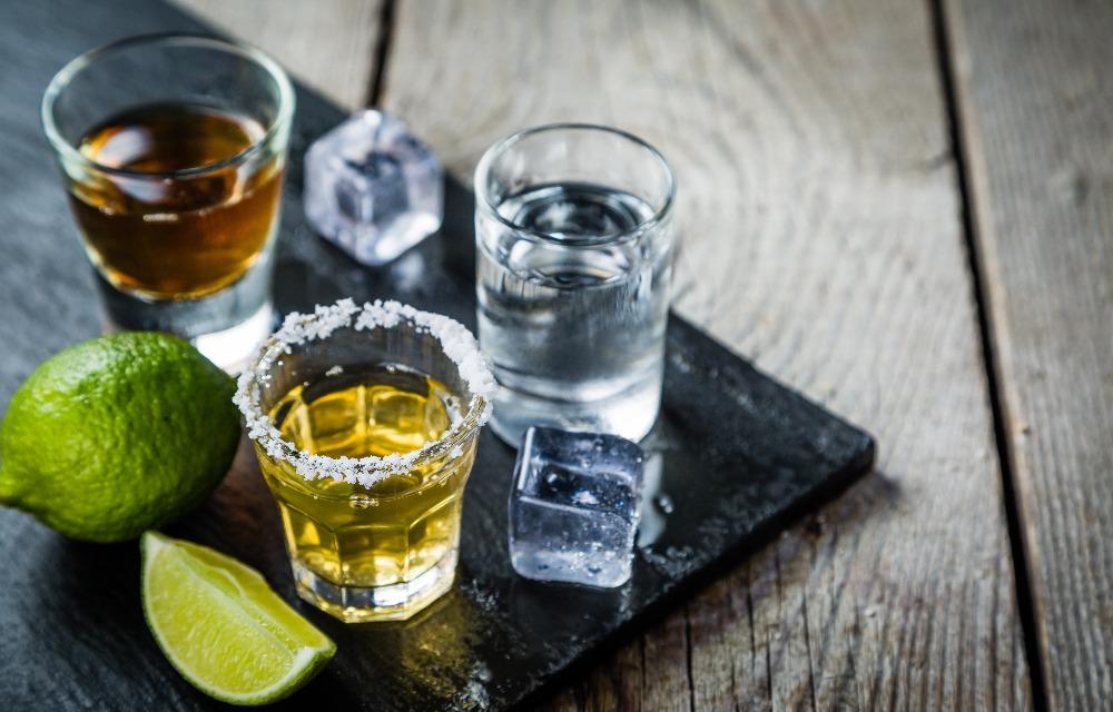 Slate with three shot glasses containing the three types of Tequila with lime wedges. Explore the diverse world of Tequila and savor each variety with a touch of lime.