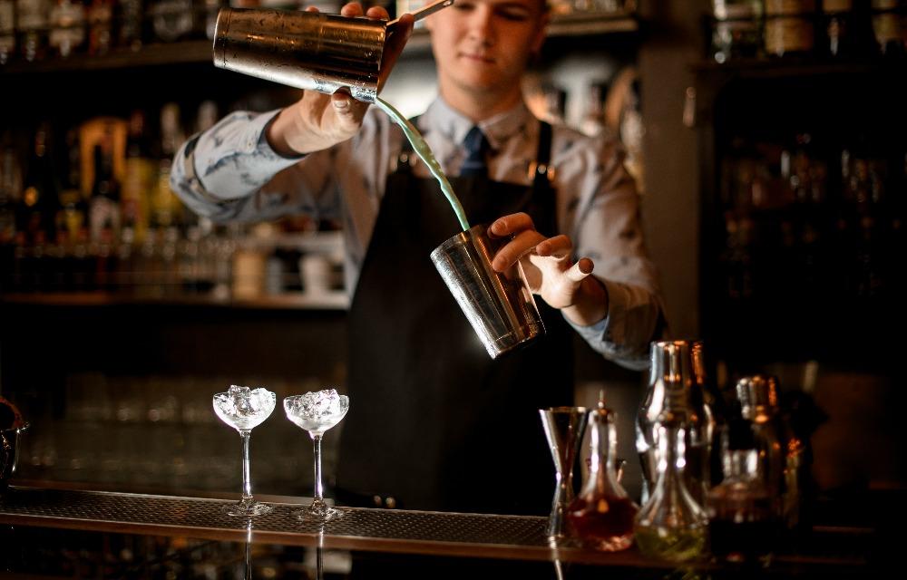 Il giovane barista sta preparando un cocktail in uno shaker. Due bicchieri con un vassoio di ghiaccio sul bancone. Scaffali con bottiglie di alcol sullo sfondo.